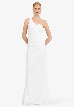 Woman wearing a white, floor-length, one-shoulder lace gown standing with hands behind her back against a plain background.