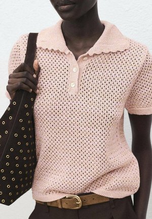 Personne à la peau foncée portant un polo tricoté rose clair, une ceinture marron et tenant un sac noir avec des œillets dorés à l'épaule.
