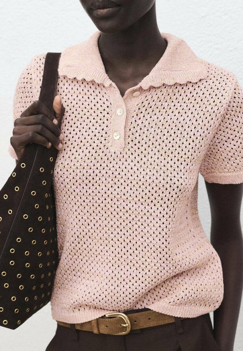 Personne à la peau foncée portant un polo tricoté rose clair, une ceinture marron et tenant un sac noir avec des œillets dorés à l'épaule.