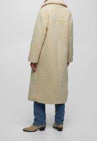 Manteau de couleur crème à texture duveteuse avec un large col, des manches longues et un ourlet longueur mi-cuisse, assorti à un jean bleu et des bottines à talons beige.