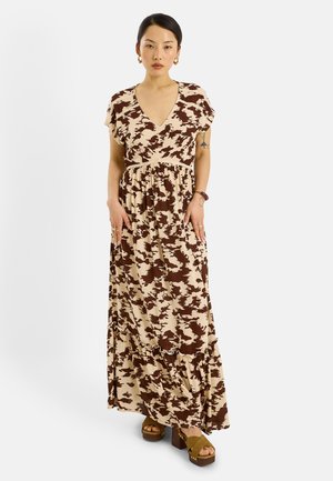 Mujer vistiendo un vestido largo con escote en V de estampado beige y marrón, con mangas cortas y sandalias de plataforma marrones, de pie frente a un fondo blanco liso.