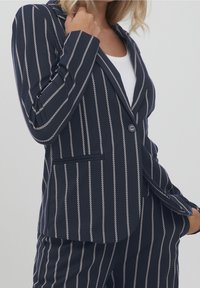 Femme portant un blazer bleu marine et un pantalon assorti à rayures blanches verticales, associés à un haut blanc en dessous.