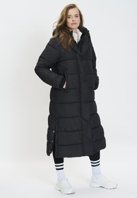 Abrigo acolchado largo negro con capucha, con diseño acolchado, bolsillos laterales y botones a presión. Lleva puesto sobre leggings negros y zapatillas blancas.
