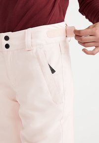 Personne attachant une patte latérale réglable sur un pantalon rose clair avec des boutons-pression noirs, portant un haut à manches longues bordeaux.