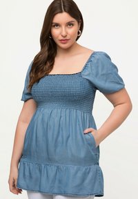 Denimoberteil mit quadratischem Ausschnitt, kurzen puffigen Ärmeln, gerafftem Oberteil und ausgestelltem Rüschensaum. Helle blaue Farbe und seitliche Taschen.