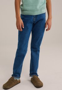 Jeans de mezclilla azul con corte recto, que cuentan con una cintura de altura media y puños doblados. Combinados con zuecos de ante marrón.