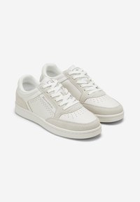 White leather sneakers with beige suede accents, perforated details, flat laces, and a rounded toe. Simple, minimalist design with a cushioned sole.