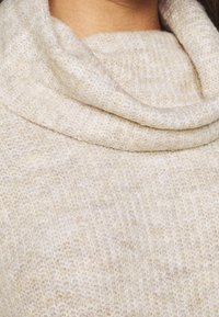 Coltrui in roomwit met een zachte, gestructureerde brei. Heeft een gedrapeerde hals en subtiele spikkels van lichtere en donkere tinten.