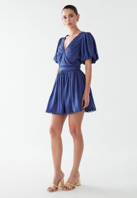 Blaues Kleid mit puffierten kurzen Ärmeln, V-Ausschnitt, gerafftem Taillenbereich und gewelltem Saum. Kombiniert mit beigen Blockabsatzsandalen.