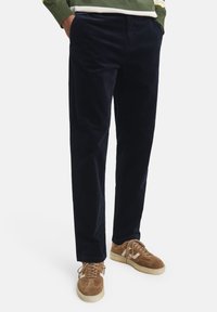 Pantaloni in corduroy blu navy con una texture liscia e taglio dritto; presentano tasche frontali e sono abbinati a sneakers in suede marrone chiaro.
