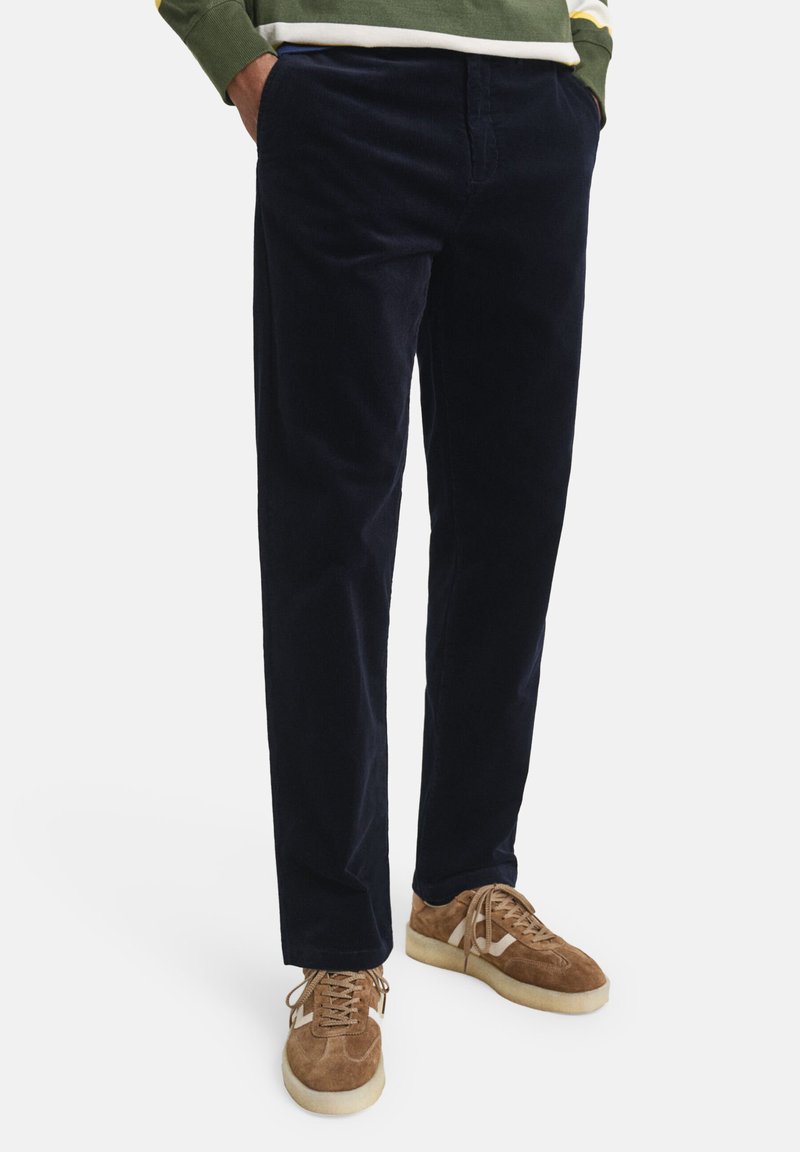 Pantaloni in corduroy blu navy con una texture liscia e taglio dritto; presentano tasche frontali e sono abbinati a sneakers in suede marrone chiaro.