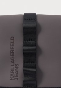 Superficie grigia opaca con nastro nero cucito verticalmente al centro e testo nero "KARL LAGERFELD JEANS" allineato verticalmente a sinistra.