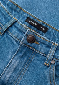 Jean en denim bleu clair avec couture orange. Présente une étiquette noire avec "ombre" et la taille M32, ainsi qu'un bouton en métal avec des détails embossés.