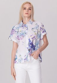 Chemise à manches courtes boutonnée de couleur blanche avec un motif fleuri aux teintes de bleu et de violet. Fabriquée en tissu léger, avec des poignets retroussés.