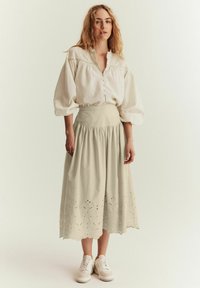 Beige, knälång kjol med scallopkant och broderade detaljer, i kombination med en löst sittande, texturerad vit blus och vita sneakers.