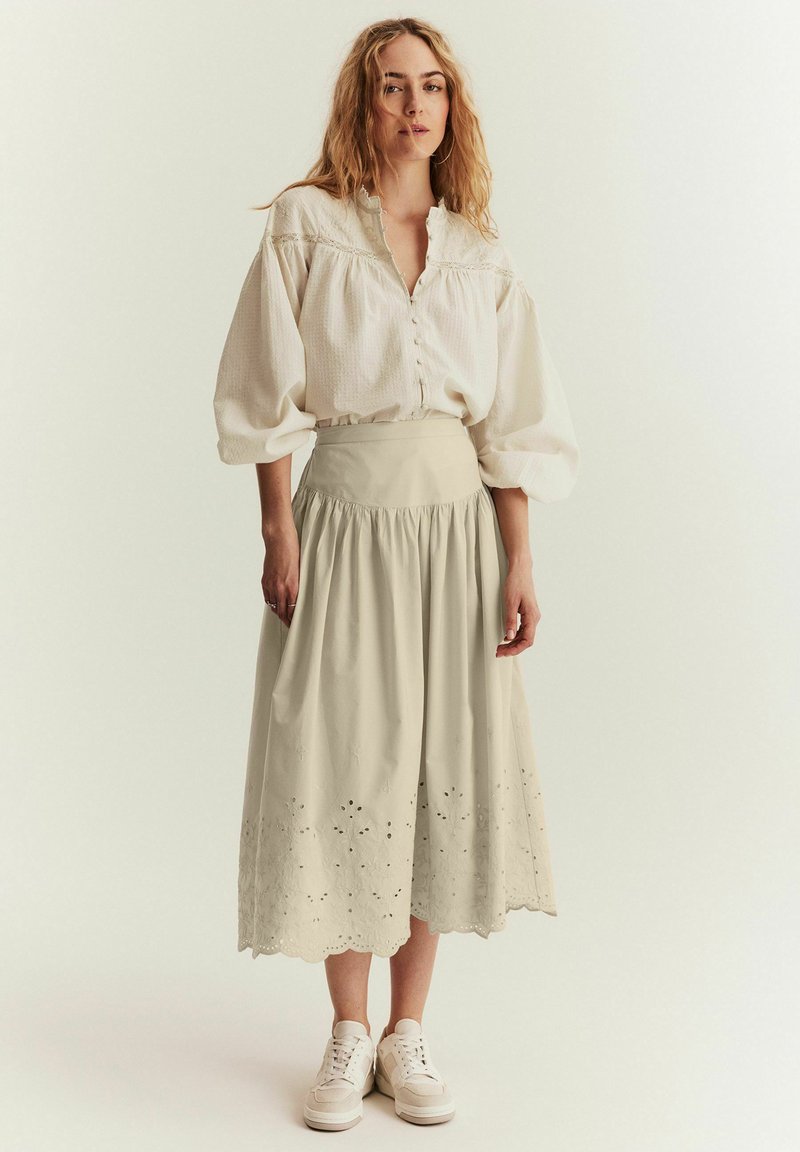Beige, knälång kjol med scallopkant och broderade detaljer, i kombination med en löst sittande, texturerad vit blus och vita sneakers.