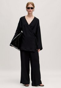 Schwarzes Wickeltop und weit geschnittene Hose aus strukturiertem Stoff. Das Model hält eine schwarze glänzende Tasche und trägt kleine schwarze Sonnenbrillen. Glatte Textur insgesamt.
