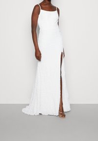 Kvinna iklädd en vit golvlång klänning med tunna axelband, texturerad tyg och en hög slits ovanför låret, tillsammans med öppna klackskor.