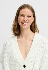 Cardigan bianco lavorato a maglia con scollo a V, bordi a costine e un singolo bottone nero sul davanti. Il tessuto appare leggero e attraente.