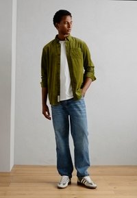 Cămașă din corduroy verde peste un tricou alb, asortată cu blugi albaștri deschiși și adidași gri cu accente verzi. Stând pe podea din lemn.