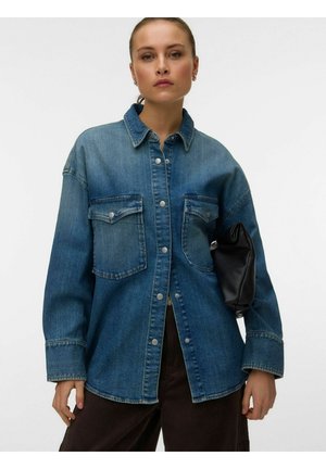 Femme portant une veste en jean bleue oversize avec des boutons-pression, tenant une pochette noire sous le bras sur un fond blanc uni.