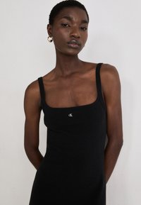 Black fitted tank top with a square neckline, thin straps, and a small white logo on the chest. Textured fabric.