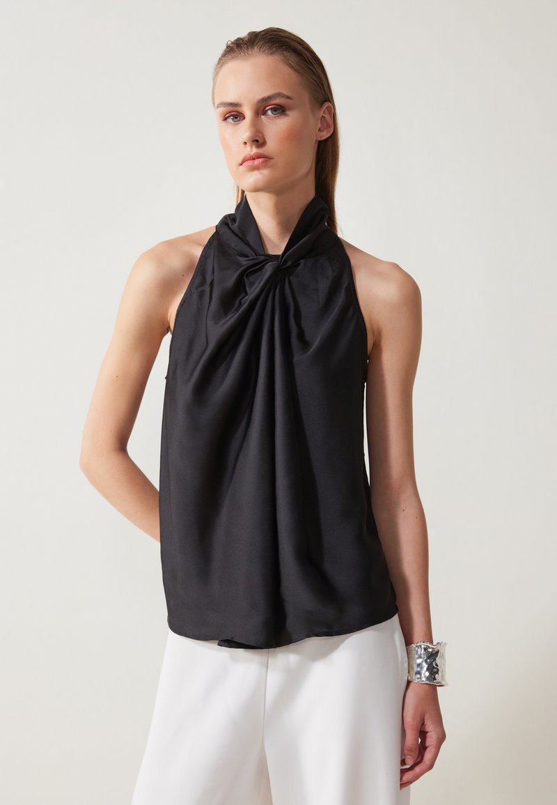 Top black a collo alto realizzato in tessuto liscio, con un dettaglio annodato arricciato all'collo. Abbinato a pantaloni bianchi e un bracciale in argento.