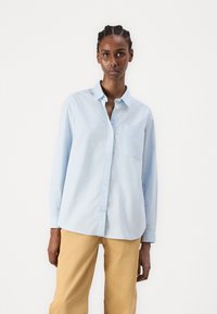 Camicia azzurro chiaro a maniche lunghe con bottoni, realizzata in tessuto morbido, dotata di colletto e di una tasca sul petto, abbinata a pantaloni beige.