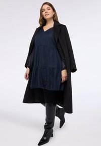 Granatowa sukienka warstwowa z dekoltem w serek i przezroczystymi długimi rękawami, zestawiona z czarnym długim płaszczem i czarnymi, sięgającymi kolan, spiczastymi kozakami.