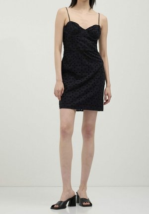 Femme portant une mini-robe noire ajustée avec des bretelles fines et des talons blocs noirs ouverts à l'avant, debout devant un fond uni.