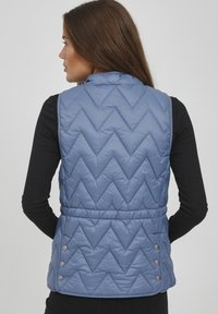 Blicksäker blå väst med zigzag-mönster, hög krage och sidoknappar. Slät textur, figursydd design och ärmlös stil.