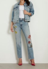 Chaqueta de mezclilla azul claro con herrajes plateados sobre una blusa blanca, combinada con jeans azul claro que presentan bordados florales en amarillo y rosa.