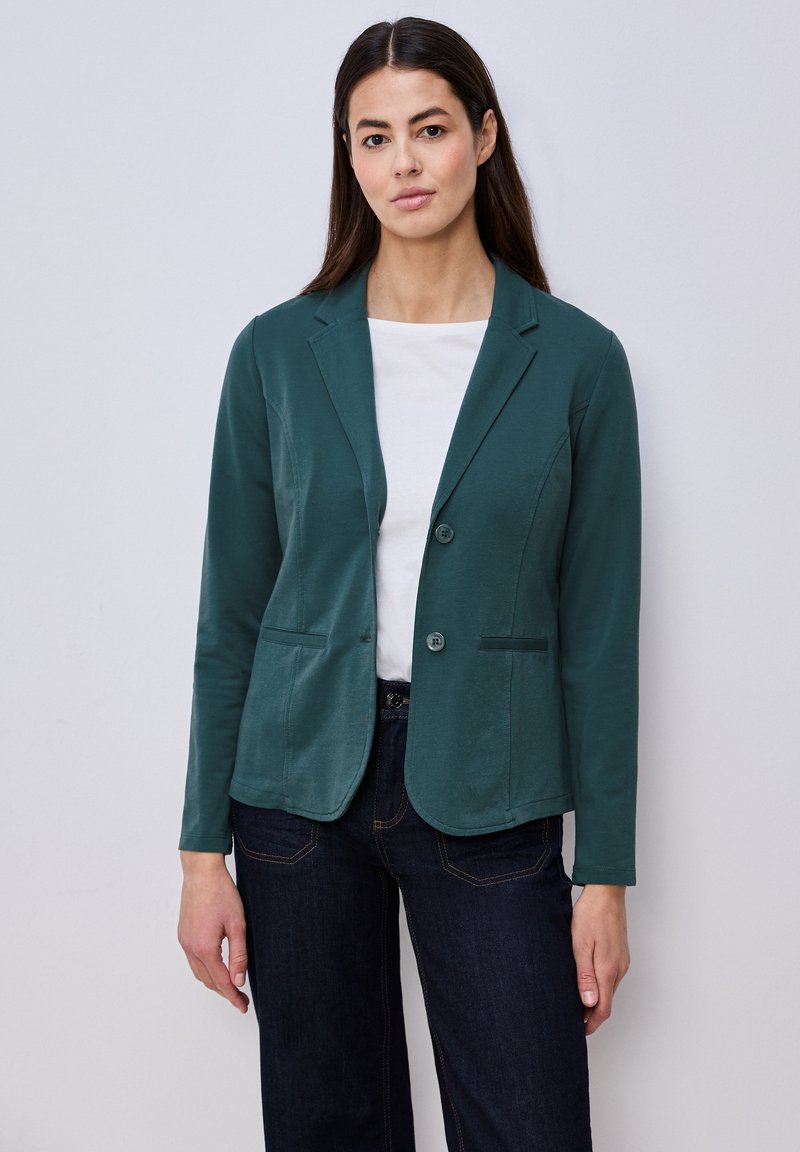 Femme portant un blazer vert foncé boutonné sur un haut blanc et un jean foncé, debout devant un fond uni gris clair.