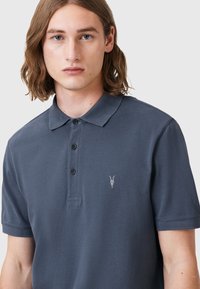 Mörkgrå pikétröja i texturerad tyg med en treknapplåsning och en liten broderad getskalle-logotyp på bröstet.