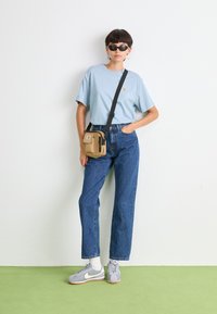 T-shirt bleu clair, jean bleu taille haute, baskets grises avec accents blancs et sac à bandoulière beige. Les accessoires comprennent des lunettes de soleil noires.