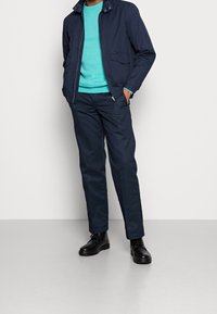 Marinblå jacka med krage och fickor, kombinerad med en turkos tröja och marinblå byxor. Svarta stövlar fulländar outfiten.