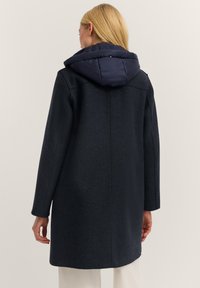 Marineblauer Mantel aus einem strukturierten Wollmix, mit einer gepolsterten Kapuze, langen Ärmeln und geradem Schnitt. Dezente Nähte und minimalistische Designelemente.