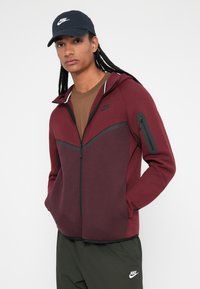 Fato de treino com fecho marrom com detalhes em preto e um bolso à frente, combinado com uma t-shirt castanha e calças verde-escuras. Logotipo da Nike visível na roupa.