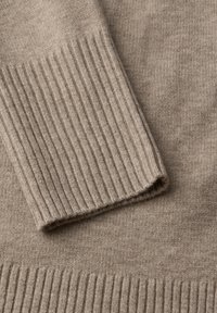 Detail béžového pleteného svetru, který se vyznačuje měkkou texturou a žebrovými manžetami a lemem. Design zahrnuje hladké a žebrované vzory.