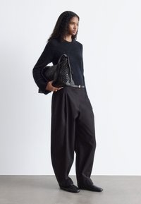 Marineblau gestrickter Pullover, weite schwarze Hosen mit Falten und eine strukturierte schwarze Handtasche. Das Modell trägt schwarze Slipper, die Haare in lockeren Locken.