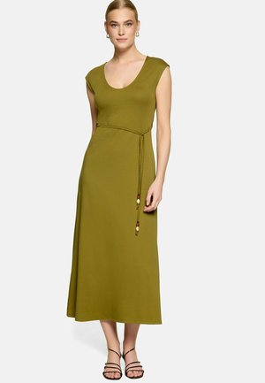 Femme portant une robe midi sans manches vert olive avec un décolleté arrondi et une fine ceinture à la taille, debout avec une main détendue sur le côté.