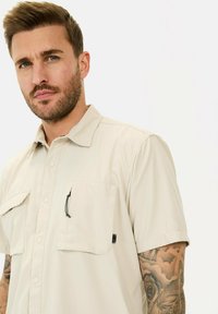 Beige kurzärmliges Hemd mit zwei Taschen vorne und sichtbaren Nähdetails. Der Stoff wirkt leicht und strukturiert.