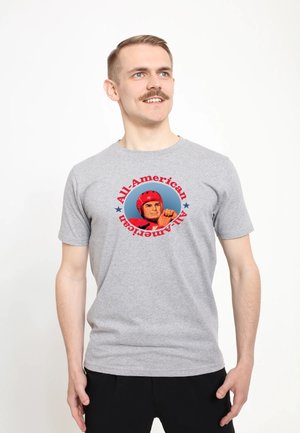 Man met snor die een grijs T-shirt draagt met een illustratie van een vintage hockeyspeler en de tekst "All-American" in rood en blauw.