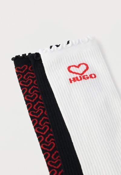 Trois paires de chaussettes côtelées : une noire avec un motif de cœurs rouges, une blanche avec le logo "HUGO" rouge, et une noire unie avec un bord supérieur volanté.
