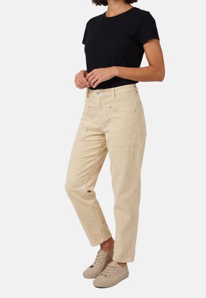Mujer con pantalones beige, camiseta negra de manga corta y zapatillas beige, de pie con las manos entrelazadas a la altura de la cintura contra un fondo blanco.