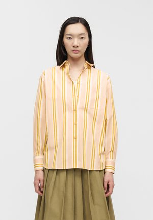 Vrouw die een perzikkleurig shirt met lange mouwen draagt met gele verticale strepen en een olijfgroene geplooide rok, staand tegen een effen achtergrond.