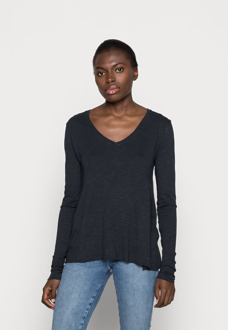 Camisa de manga longa, com decote em V na cor navy, feita de um tecido leve, apresentando um corte descontraído e uma bainha ligeiramente curvada. Usada com jeans azuis.