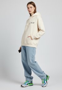 Felpa crema con tasca frontale e cappuccio con coulisse, abbinata a jeans aderenti azzurri e scarpe da corsa multicolori.