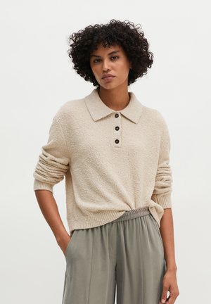 Femme aux cheveux bouclés portant un pull polo beige texturé à manches longues et un pantalon gris ample, les mains dans les poches, sur un fond uni.