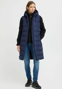 Lang marineblå polstret vest med hette, med glidelås og sidelommer, brukt over en svart genser og blå jeans, kombinert med svarte sko.